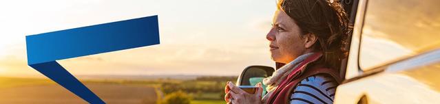 Woman with a scarf around her neck leaning on her car and holding a cup of hot beverage on the side of a country road.